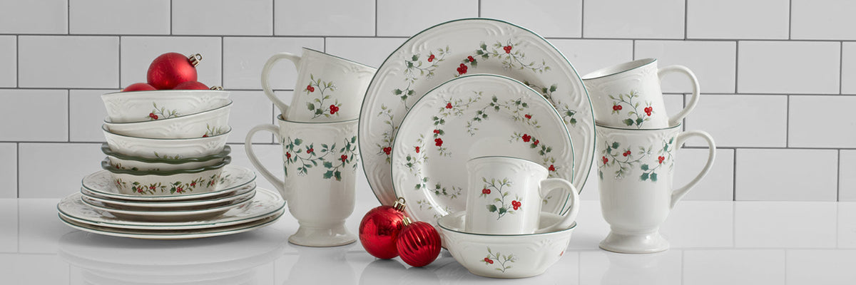 Winterberry white ceramic dinnerware with holly and red berries, featuring stacked plates, bowls, mugs and a cup against a white tile backdrop.