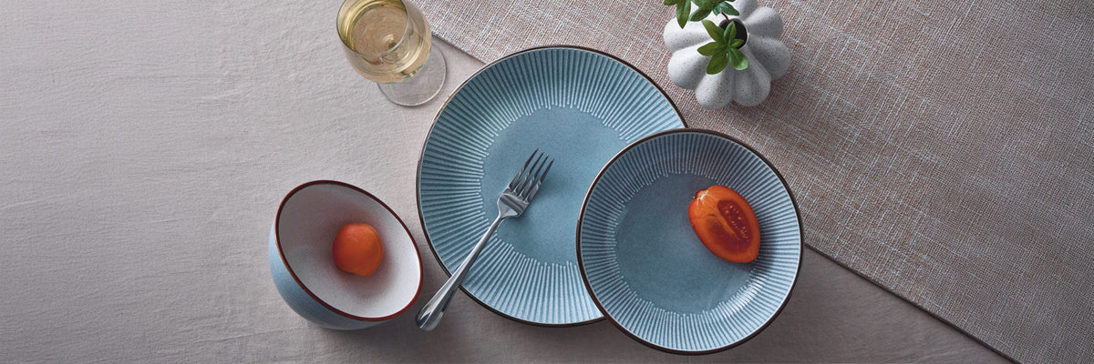 Top view of the Arlie blue ribbed dinnerware set with a large plate and a smaller plate, fork, a bowl with an orange fruit, a halved orange fruit on the other plate, a wine glass, and a small plant on a textured surface.