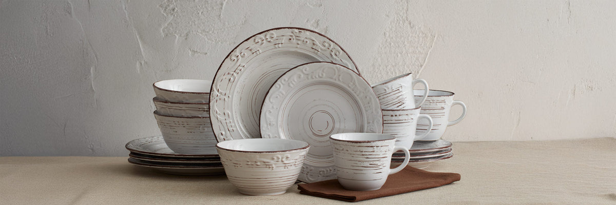 Trellis Full Collection white ceramic dinnerware arranged on a beige table with plates bowls and mugs featuring brown distressed glaze