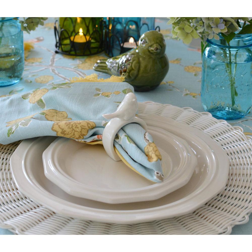 Table setting with layered Heritage Dinner Plates a light blue napkin in a white napkin ring on a wicker placemat and blue glass jars